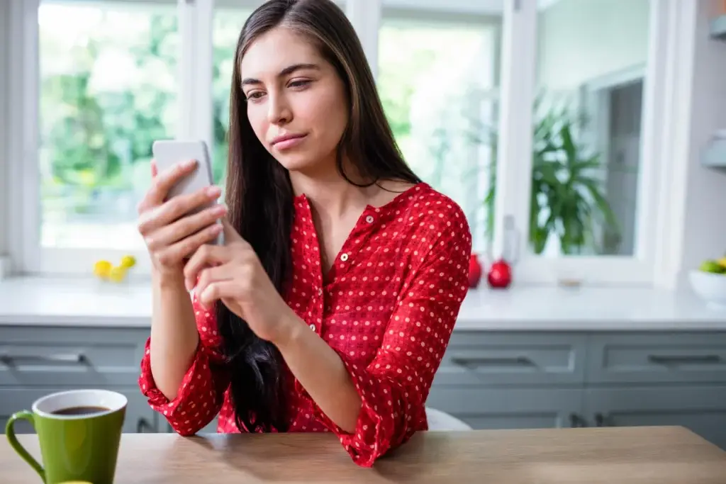 A woman sitting at her kitchen table scrolling on her phone with a cup of coffee.