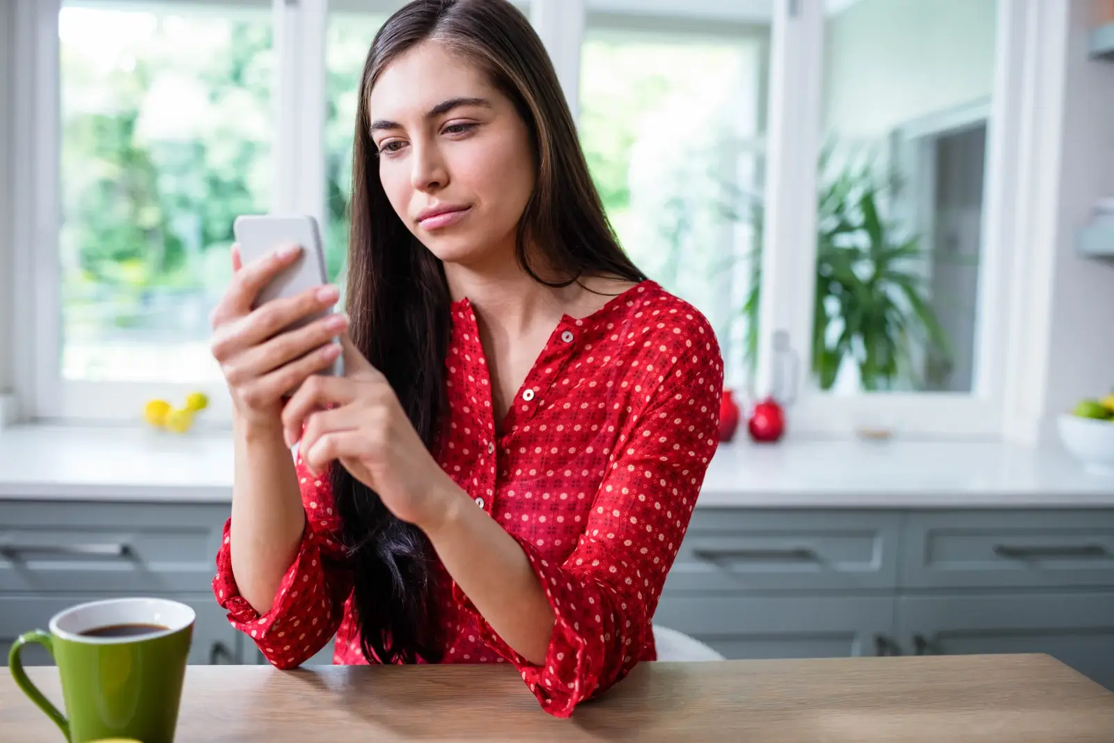 A woman sitting at her kitchen table scrolling on her phone with a cup of coffee.