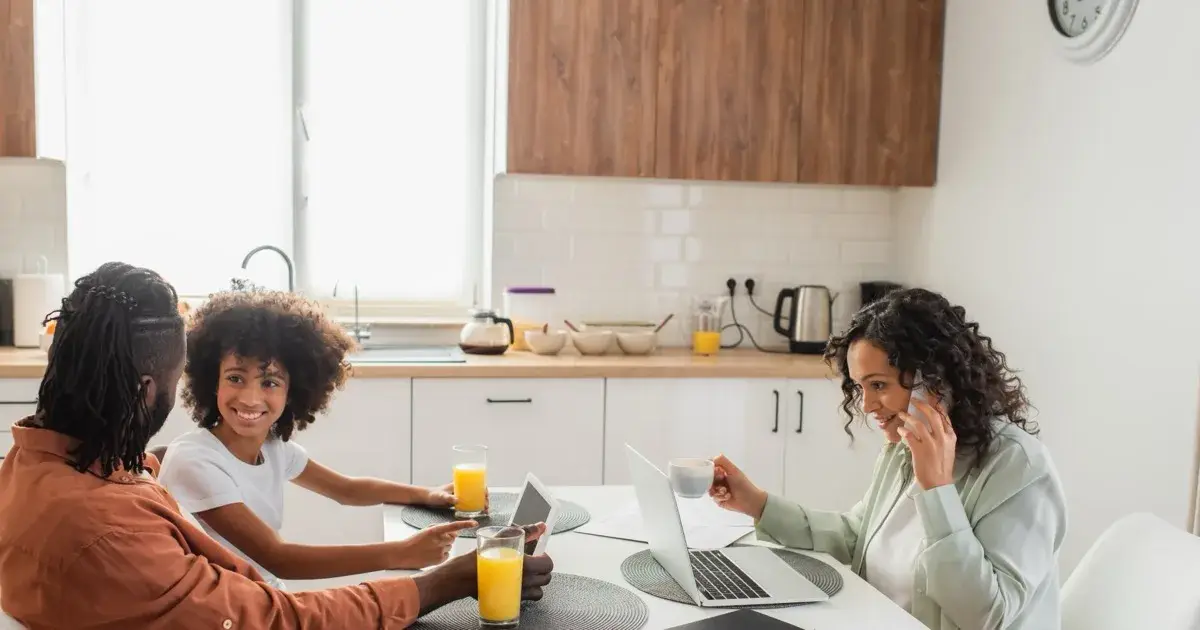 A family using phones, a tablet and a laptop together at a kitchen table.