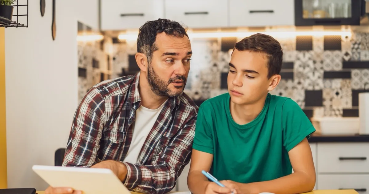 A teenager working at a kitchen table with a parent nearby.