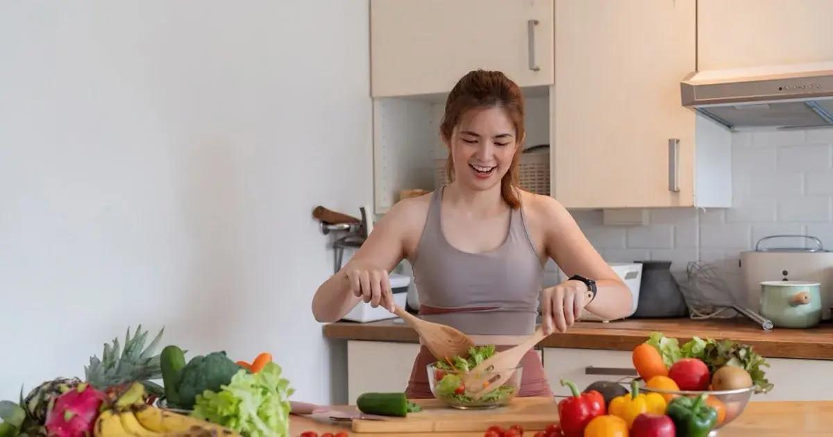 A woman preparing a salad at a kitchen counter surrounded by fresh vegetables and fruit.