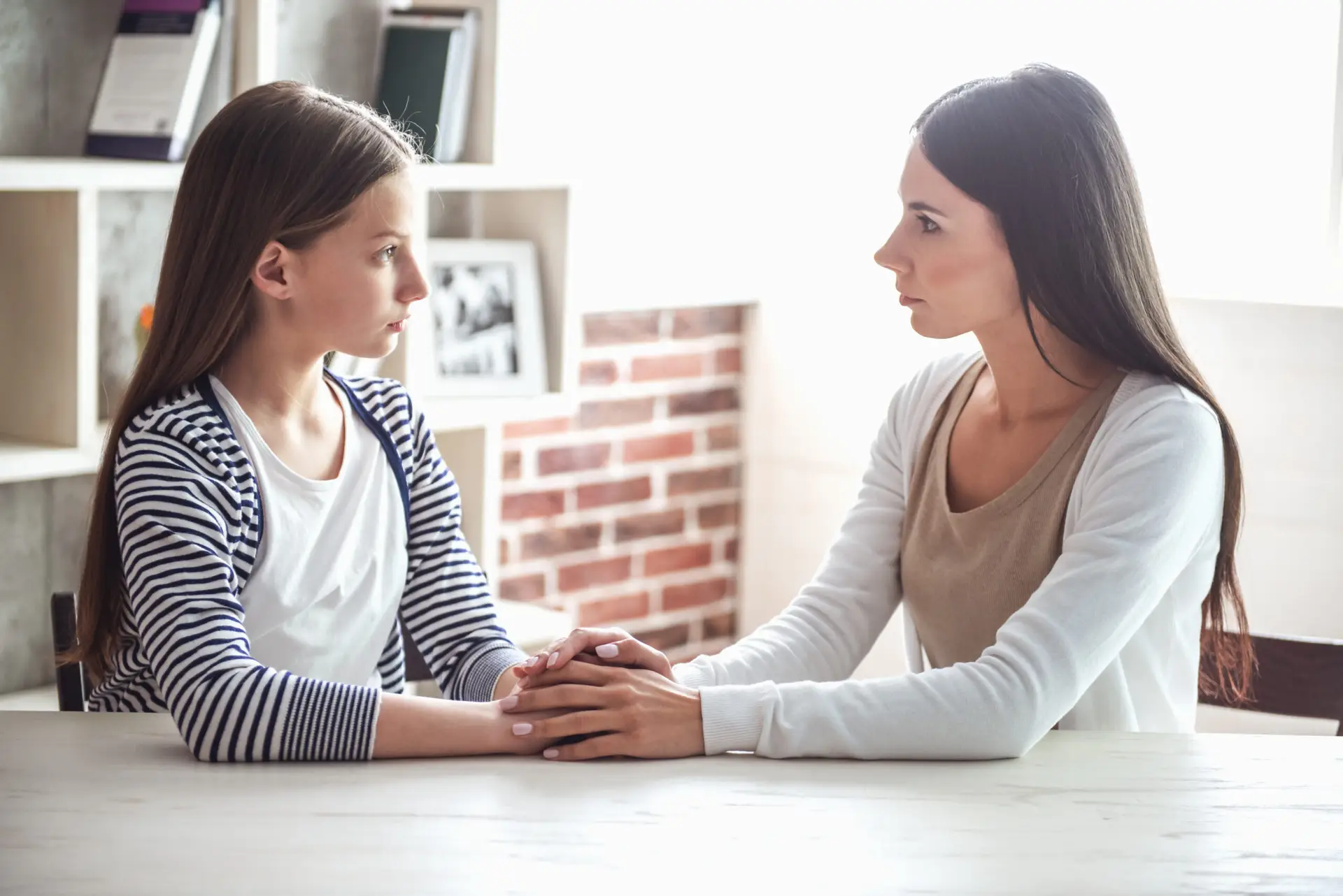 A mother and her teenage daughter sit across a table holding hands during a calm, serious conversation.
